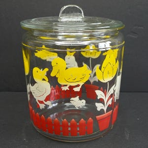 May include: A clear glass jar with a matching lid. The jar is decorated with yellow ducks, white rabbits, red picket fences, and yellow tulips. The jar is a vintage kitchen storage container.