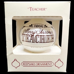 May include: A white glass Christmas ornament with the text "Merry Christmas, Teacher" in brown lettering. The ornament is in a white box with the text "Teacher" and "Keepsake Ornament" in red lettering.