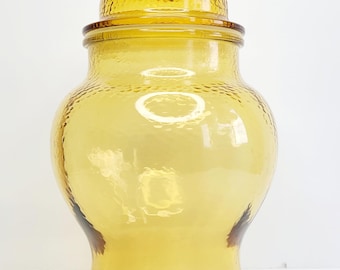 Pode incluir: Um grande frasco de boticário de vidro cor de âmbar com uma superfície texturizada e com covinhas. O frasco apresenta um corpo largo e arredondado, um pescoço curto e uma tampa removível a condizer com um puxador esférico de vidro liso.