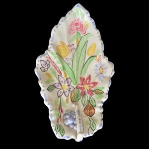 Puede incluir: Un plato de cerámica con forma de hoja con un diseño floral pintado a mano. El plato presenta una base blanca con un borde azul punteado y un ramo de flores coloridas, que incluyen rosas rosas, tulipanes amarillos y flores rojas. También se incluye un pequeño racimo de uvas azules.