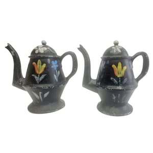 Puede incluir: Dos pequeñas teteras de metal negro con diseños florales. Cada tetera presenta un pico curvo, un asa redondeada y una tapa con un pequeño pomo. Los diseños florales incluyen tulipanes amarillos, flores azules y flores blancas, añadiendo color.