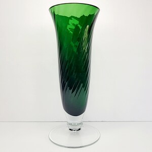 May include: A tall, green glass vase with a swirling design. The vase has a clear glass base with a wide, circular foot.