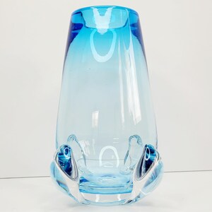 May include: A light blue glass vase with a gradient from dark blue to light blue. The vase has a unique, organic shape with a wide base and a narrow neck. The base of the vase has a series of small, rounded protrusions.