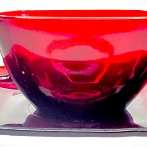 May include: A red glass teacup and saucer set. The cup and saucer are both a deep red colour and have a slight ombre effect, fading to a darker red at the bottom.