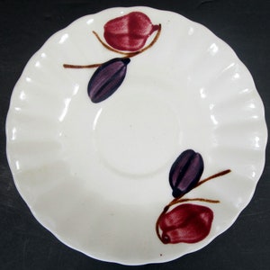 May include: White ceramic saucer with a scalloped edge and a floral design. The design features two red and purple stylized flowers with brown stems.