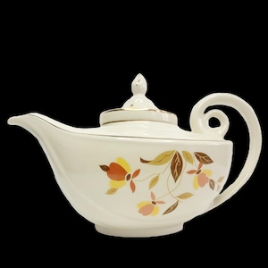 May include: Cream ceramic teapot with a curved handle and spout. The teapot has a floral design in yellow, orange, and brown. The lid is topped with a small finial and trimmed with gold. A classic tea accessory.