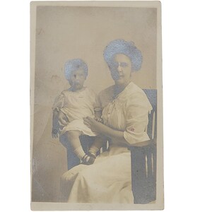 Puede incluir: Una fotografía vintage en blanco y negro de una mujer sosteniendo a un niño pequeño en su regazo. La mujer lleva un vestido blanco y un sombrero. El niño lleva un vestido blanco y tiene el pelo corto.