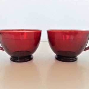 Puede incluir: Dos tazas de vidrio rojo con asas. Las tazas son redondas y tienen un ligero borde alrededor de la parte superior. Las tazas están sobre una superficie blanca.