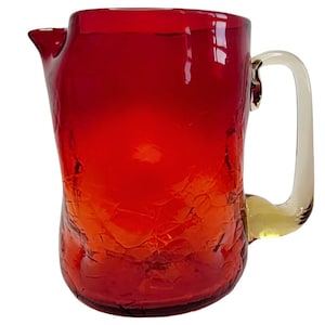 May include: A vibrant red glass pitcher with a textured surface and a clear handle. The pitcher has a spout for easy pouring and is likely used for serving beverages. The glass has a crackled appearance, adding to its unique aesthetic.