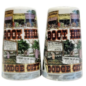 May include: Two ceramic salt and pepper shakers with a vintage design. The shakers feature illustrations of a town with the text "Boot Hill" and "Dodge City". The design includes text that reads "Shoot-Em-Up Jake".