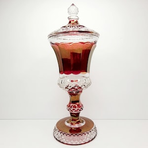 Puede incluir: Un gran plato de dulces de vidrio transparente con un acabado rojo iridiscente. El plato tiene una base de pedestal y una tapa con un pomo.