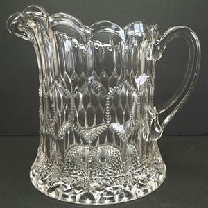 May include: A clear glass pitcher with a scalloped rim and a diamond-shaped pattern. The pitcher has a handle and a wide base.