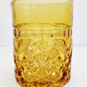 May include: An amber-coloured glass tumbler with a geometric diamond pattern. The glass has a smooth rim and a warm, golden hue. The design is reminiscent of mid-century glassware. The tumbler is suitable for serving beverages. The height is approximately 10 cm.