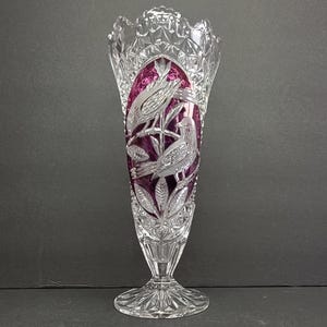 Puede incluir: Un jarrón de cristal transparente con una parte superior acampanada y una base de pedestal. El jarrón presenta un panel central de color púrpura intenso con pájaros y follaje grabados. El cristal está tallado intrincadamente con patrones geométricos.