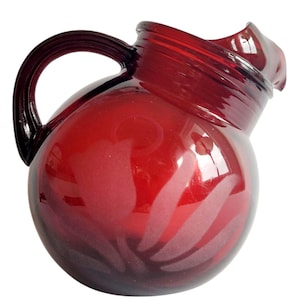 Puede incluir: Una jarra de vidrio rojo rubí con cuerpo redondeado y asa curva. La jarra tiene un pico inclinado único y un sutil patrón de hojas grabado en el vidrio. Es un artículo decorativo.