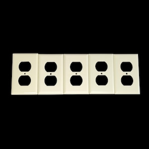 Puede incluir: Cinco cubiertas de tomacorrientes de color blanquecino dispuestas en fila sobre un fondo negro. Cada cubierta tiene dos aberturas ovaladas para enchufes y una pequeña abertura circular encima.