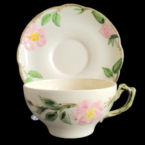 Könnte beinhalten: Ein Vintage-Teetassen- und Untertassen-Set mit einem Blumendesign. Tasse und Untertasse sind beide cremefarben mit einem Muster aus rosa Rosen und grünen Blättern.