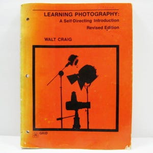 May include: An orange book titled "Learning Photography: A Self-Directing Introduction, Revised Edition" by Walt Craig. The cover features a black silhouette of studio lighting equipment. The book has three holes on the left side.