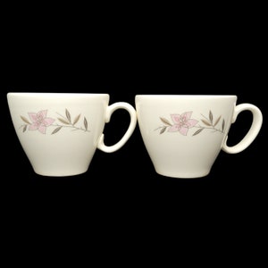 May include: Two white ceramic teacups with a floral design. The cups have a pink flower with brown stems and leaves.