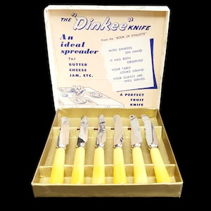 Pode incluir: Um conjunto vintage de seis espalhadores "Dinkee Knife" numa caixa dourada. As facas têm cabos amarelos e lâminas prateadas com gravuras decorativas. A tampa da caixa apresenta o texto "Um espalhador ideal para manteiga, queijo, geleia, etc."