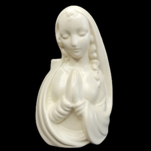 Op de afbeelding: Crèmekleurige keramische buste van een vrouw met haar handen gevouwen in gebed. De figuur draagt een gedrapeerde sluier en heeft een serene uitdrukking. De buste staat tegen een zwarte achtergrond.