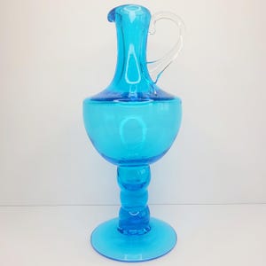 May include: A tall, turquoise-colored glass pitcher with a clear handle. The pitcher has a bulbous body, a slender neck, and a flared base. The handle is arched and attached to the neck and body. The pitcher is likely used for serving beverages.