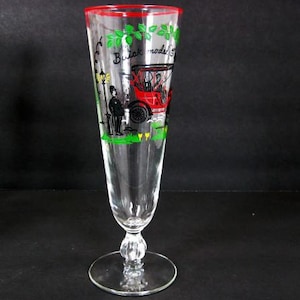 Puede incluir: Una copa de champán de cristal transparente con borde rojo y base decorativa. La copa está adornada con una colorida ilustración de un coche rojo vintage, una figura y el texto "Buick model 5". El follaje verde acentúa la parte superior.