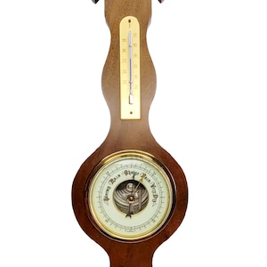 May include: A wall-mounted wooden weather station featuring a vertical thermometer with a brass scale, a central circular barometer with a white dial marked Stormy, Rain, Change, Fair, and Very Dry, and a small brass circular hygrometer at the base.