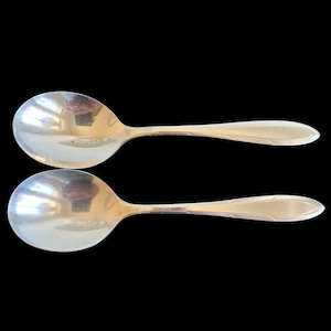 May include: Two silver soup spoons with oval bowls and elongated handles. The spoons have a reflective surface and a decorative design near the handle's base. The background is solid black.