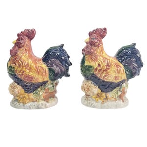 Può includere: Due statuette di gallo in ceramica. I galli hanno creste rosse, becchi gialli e piumaggio colorato nelle tonalità del giallo, arancione, verde e blu. Sono appollaiati su una base con dettagli floreali e di foglie.