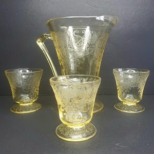 Op de afbeelding: Een vintage gele glazen kan met een handvat en drie bijpassende voetglazen. Het glaswerk heeft een geëtst bloemen- en vlinderontwerp. De set wordt op een donkere ondergrond gepresenteerd.