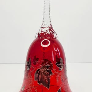 May include: A ruby red Bohemian crystal bell with a clear, twisted handle. The bell features a silver grape leaf design and a silver rim. A small, white label is affixed to the bell.