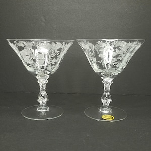 May include: Two clear glass stemmed cocktail glasses with a floral etched design. The glasses have a wide bowl and a delicate stem with a decorative knob.