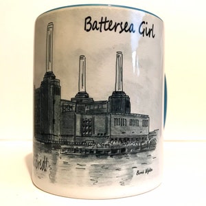 May include: A white ceramic mug with a blue rim features a black and white illustration of Battersea Power Station in London. The text "Battersea Girl" is written in black on the mug. The artist's signature "Bernie Wigston" is written in black at the bottom of the mug.