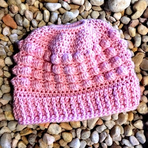 May include: A pink crocheted beanie hat with a textured, bumpy design. The hat has a folded brim.