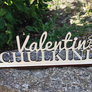 Könnte beinhalten: Holzschild mit dem Namen "Valentin" in Schreibschrift über dem Wort "SCHULKIND" in Großbuchstaben. Das Schild hat ein Kuchendesign auf der rechten Seite. Das Schild ist aus hellem Holz mit einem dunkelbraunen Sockel gefertigt.