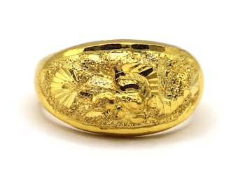 May include: A gold-colored ring with a detailed dragon design. The ring has a textured surface and a rounded shape. The dragon is the central focus, with intricate carvings and a sunburst pattern.