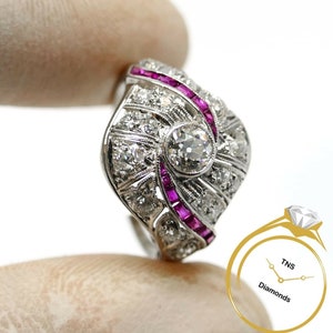 May include: A vintage-style ring with a large round diamond surrounded by smaller diamonds and a swirl of pink rubies. The ring is set in white gold.