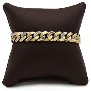 May include: A gold and silver-toned curb chain bracelet. The bracelet is displayed on a brown leather cushion. The chain links alternate between gold and silver tones. The bracelet is a piece of jewellery.