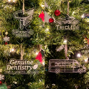 Op de afbeelding: Vier heldere acryl kerstversieringen die aan een groene boom hangen. De ornamenten tonen de tekst "GROUNDED WELLNESS", een motorfiets met de tekst "TRICLED", "Genuine Dentistry" en een busje met de tekst "FEAST & FETTLE".