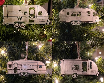 May include: Four silver-colored camper ornaments hanging on a Christmas tree. Each ornament is a different camper design with family names and the year 2023 etched into the metal. The ornaments are suspended by twine.