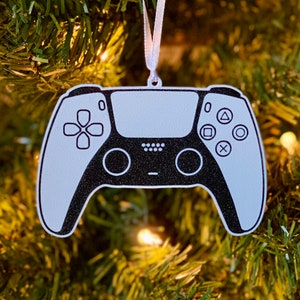 May include: White and black plastic Christmas bauble shaped like a video game controller. The controller has buttons, a directional pad, and two joysticks.