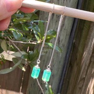 Puede incluir: Un par de pendientes de cadena plateada con cuentas cilíndricas de color verde brillante. Los pendientes cuelgan de una clavija de madera sobre un fondo de follaje verde y una valla de madera.