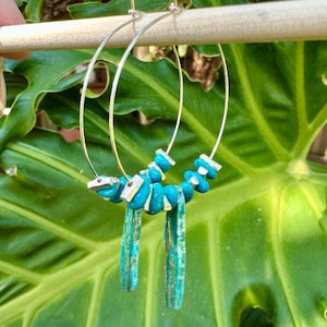 Puede incluir: Pendientes de aro plateados con cuentas turquesas y colgantes alargados de piedra color turquesa. Los pendientes se exhiben sobre un fondo de hojas verdes, mostrando los vibrantes colores y texturas de la joyería.