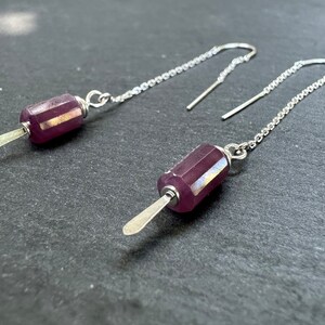 May include: A pair of silver dangle earrings with a purple gemstone pendant on each. The pendant is shaped like a popsicle and has a silver stick.