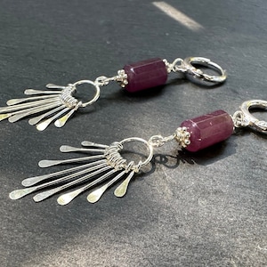 May include: A pair of silver earrings with a purple gemstone dangle. Each earring features a silver hoop and a silver fan-shaped design with multiple thin, pointed pieces.