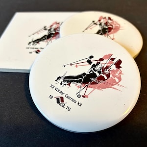 May include: White round button with a black and white illustration of a skier in a red and white background. The text "XII Winter Games XII 1976" is printed on the button.
