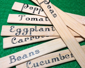 Puede incluir: Un conjunto de marcadores de plantas de madera con letras negras. Los marcadores están etiquetados con los nombres de diferentes verduras, incluyendo "Peas", "Tomatoes", "Eggplant", "Carrots", "Beans", y "Cucumber".