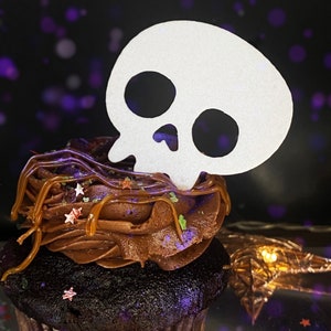 May include: A chocolate cupcake with brown frosting and sprinkles topped with a white paper skull cutout.