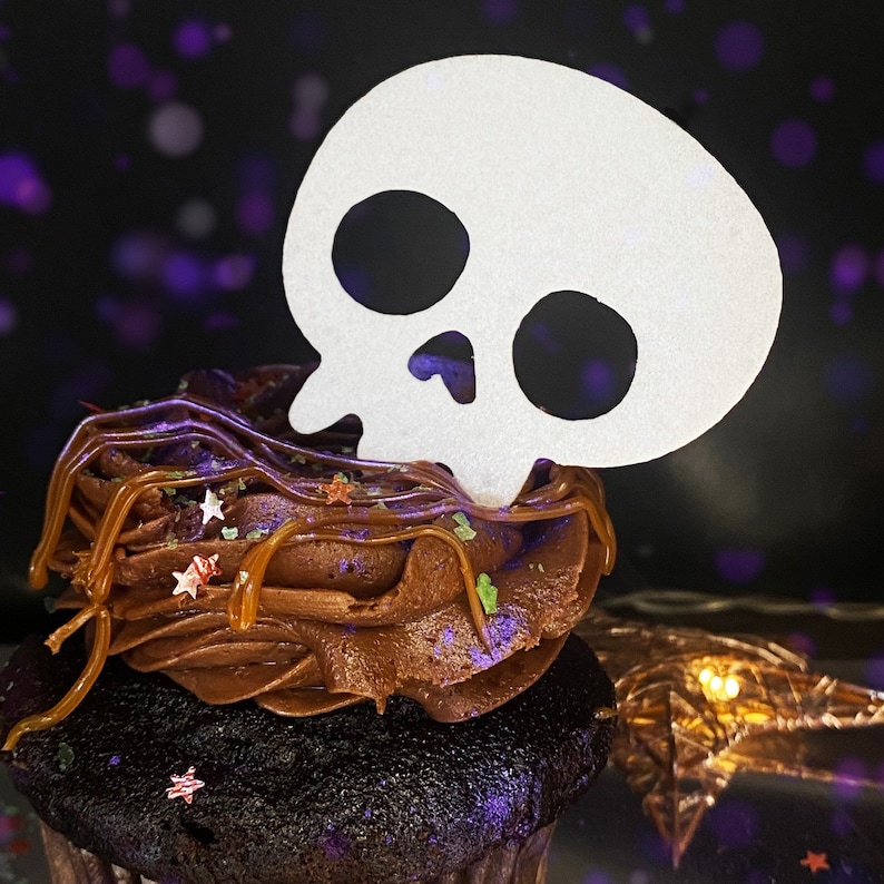 May include: A chocolate cupcake with brown frosting and sprinkles topped with a white paper skull cutout.